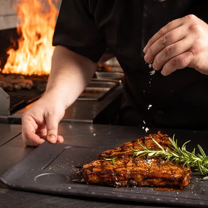 A chef seasoning a steak at the grill, flames behind